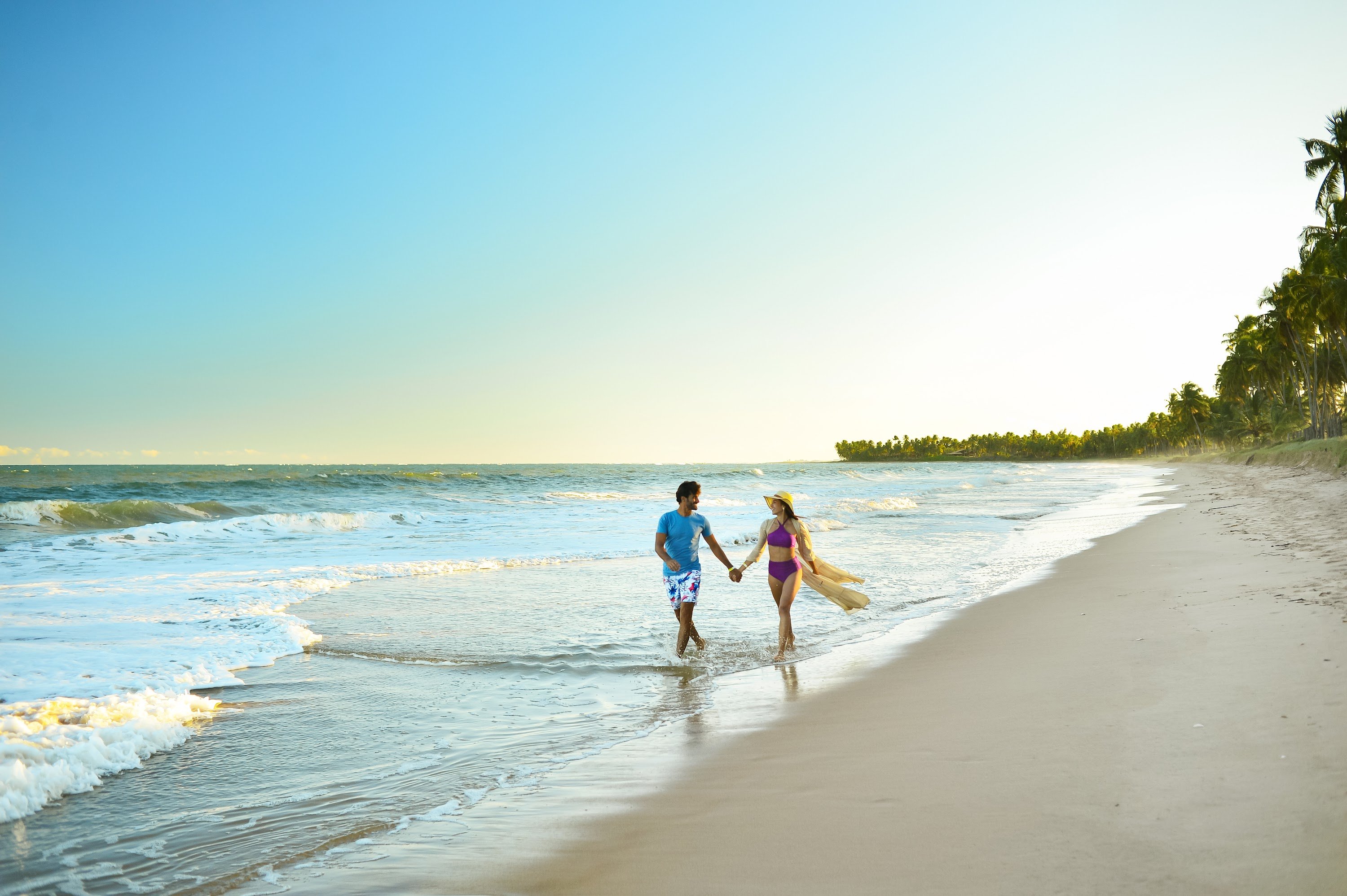 Casal caminhando sobre a areia branca, ao lado do mar e dos coqueirais da praia de Ipioca.