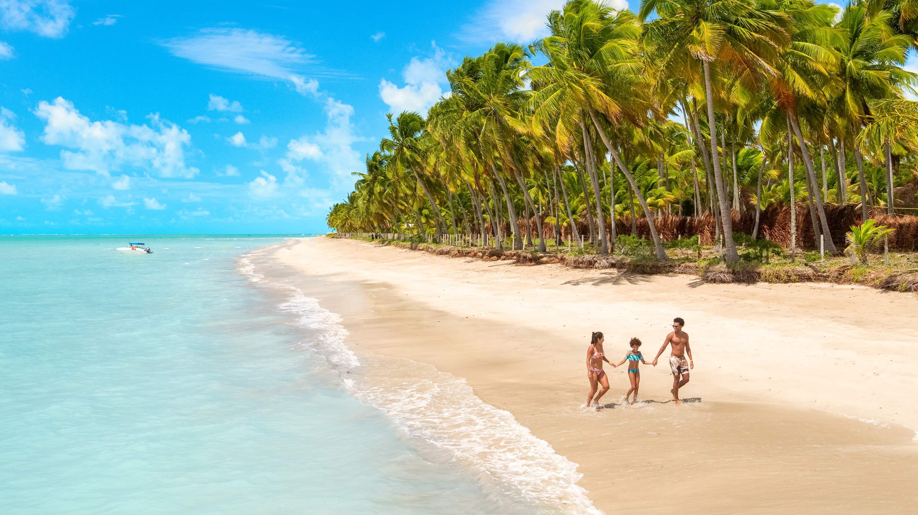 Família caminha sobre faixa de areia branca entre o mar de água azul turquesa e vasta vegetação de coqueirais na Praia de Ipioca, em Alagoas.