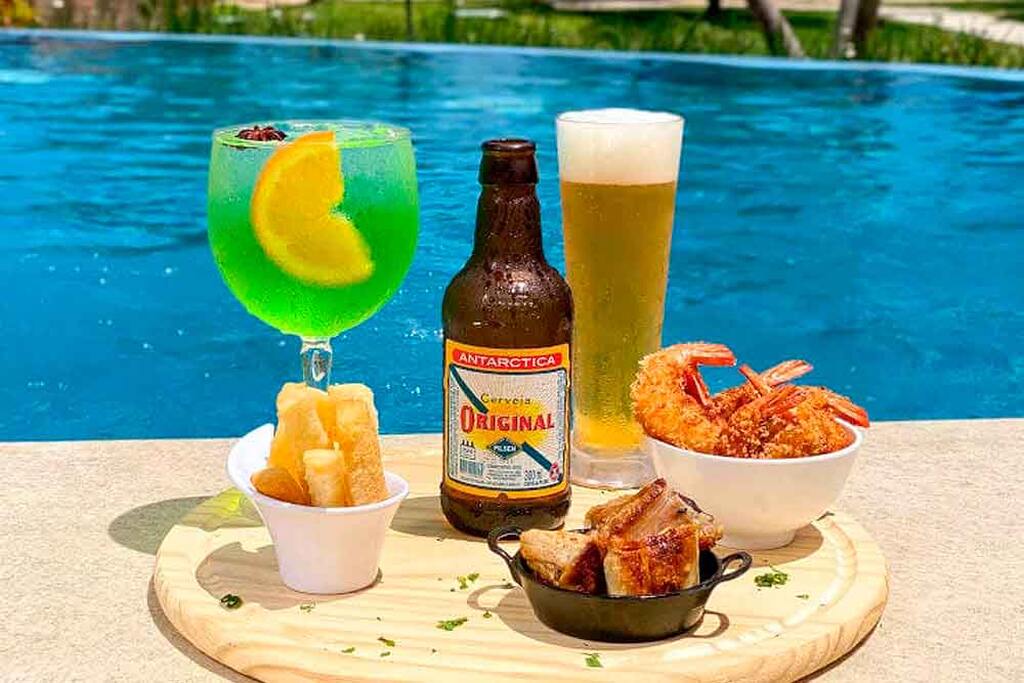 Imagem de uma bebida colorida verde com limão, garrafa de cerveja, cerveja servida no copo, mandioca e camarões fritos, vista de uma piscina azul ao fundo.