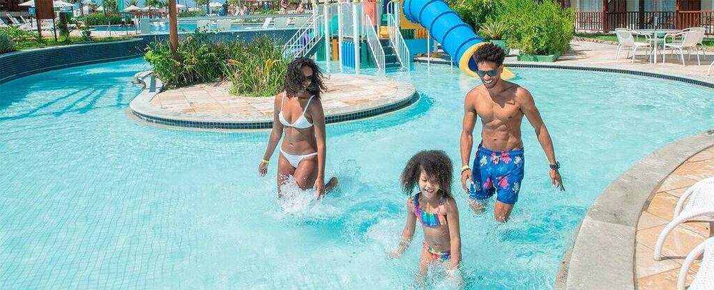 Família se divertindo e aproveitando o dia na piscina do resort Salinas ao ar livre com escorregador infantil ao fundo.
