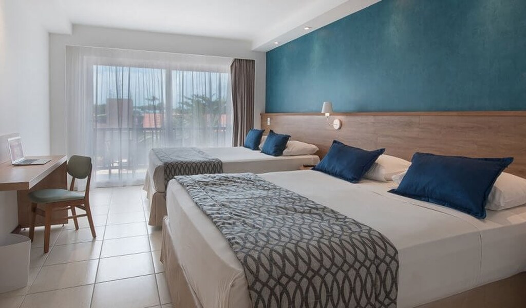 Quarto de hotel moderno com duas camas de casal, decoração clean e vista para a varanda com cortinas transparentes, ideal para viagens de lazer.