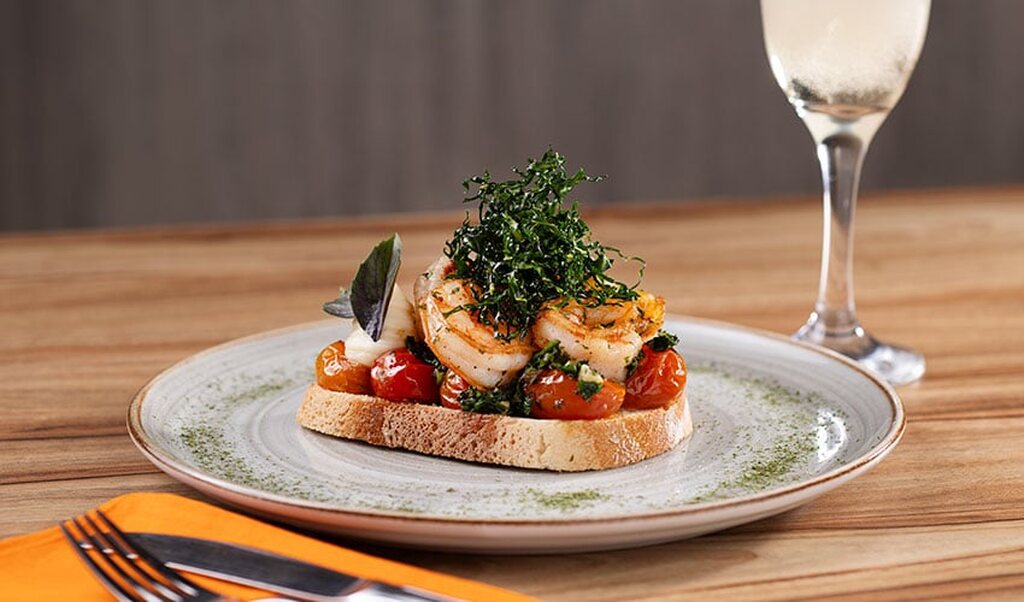 Prato sofisticado com pão, camarão, tomate-cereja, alface e vinho branco ao fundo, servido em uma mesa de madeira. Uma experiência gourmet.